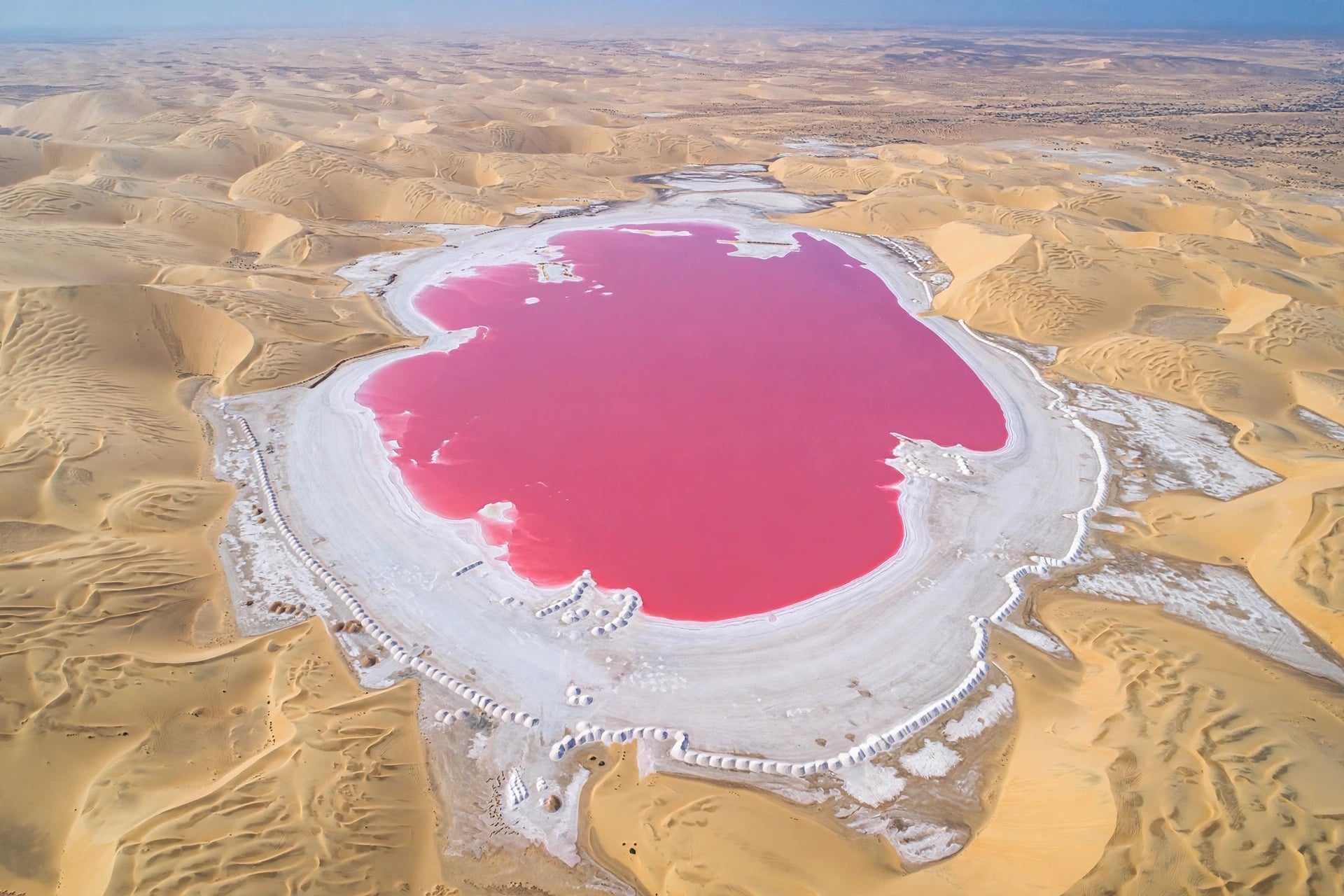 [AERIAL VIEW LAKE RETBA]  -Lake Retba (Pink Lake), Senegal - aerial drone view - Shape: Oval lake with bright pink water in center - Color: Deep pink to magenta pink water - Border: White ring around lake - salt and sand beach - Salt Piles: Hundreds of small white mounds of salt around edge of lake